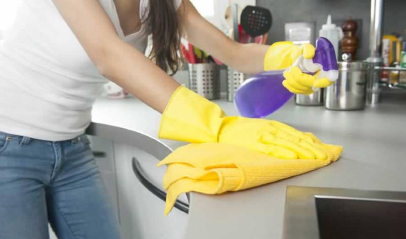 Woman in white top, jeans and yellow gloves scrubbing inside a kitchen holding spray bottle in hand