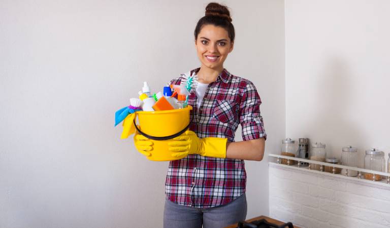 Woman in check shirt and jeans holding a yellow basket filled with tools, supplies.
