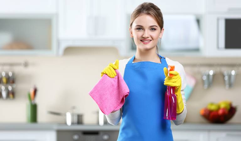 Woman in yellow gloves holding a red bottle and pink cloth standing inside a kitchen