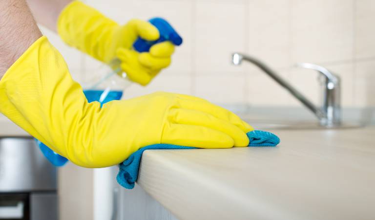 Hand in yellow gloves holding a bottle cleaning surface inside kitchen with green cloth.
