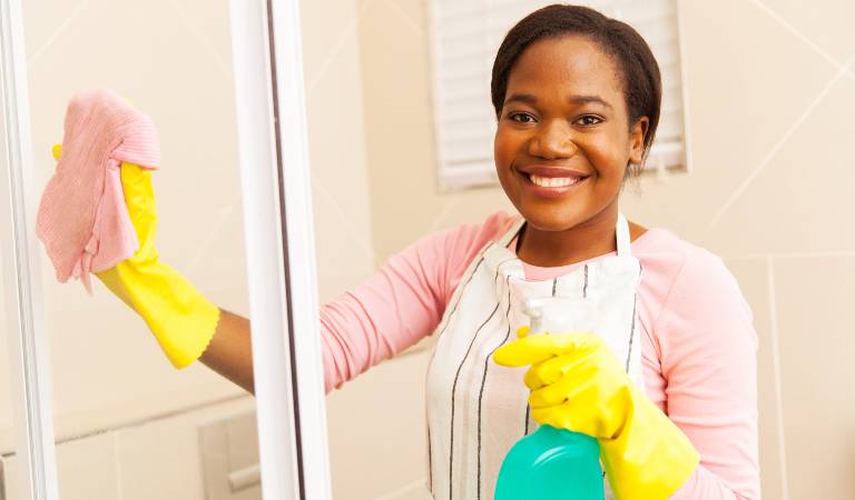 Smiling woman in yellow gloves holding a green bottle cleaning shower glass with pink cloth.
