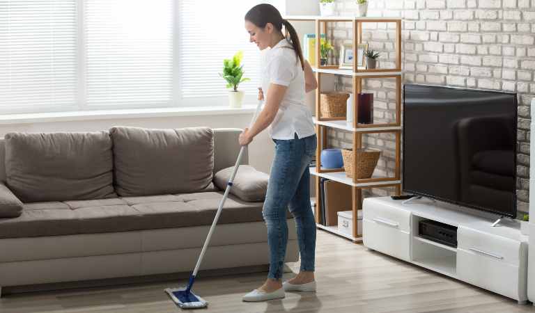 Woman in white top and blue jeans cleaning hardwood floors with a cleaning stick.