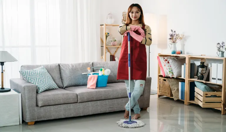 Female housekeeper standing while keeping her arms on the mop and cleaning supplies on sofa