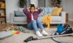 Sad tired confused black housewife hold head with hands sit on floor with vacuum or steam cleaner