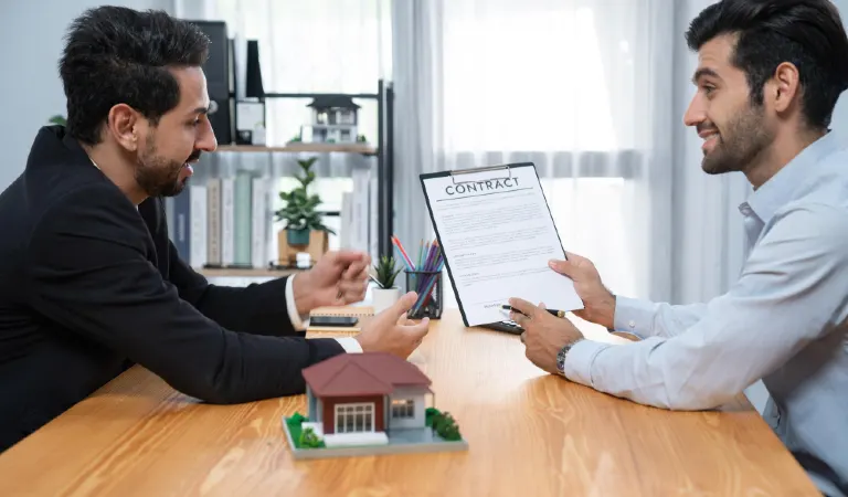 Two men sit at a desk, one of them is holding a document and house model on table