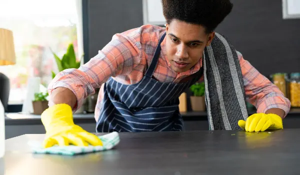 Focused biracial man wearing apron and rubber gloves cleaning countertop in home kitchen